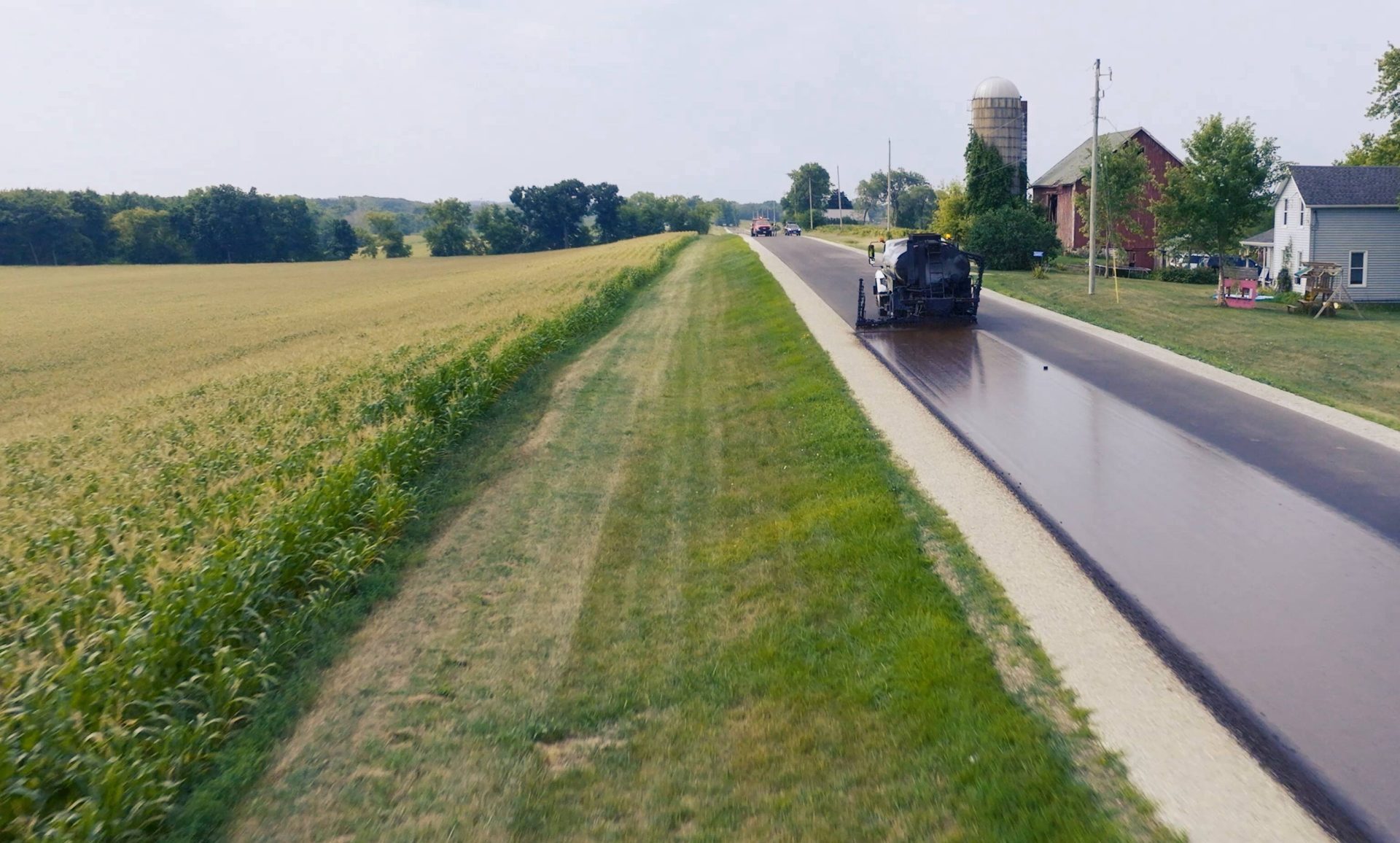 A truck drives down a country road spraying a thin asphalt emulsion across the road's surface